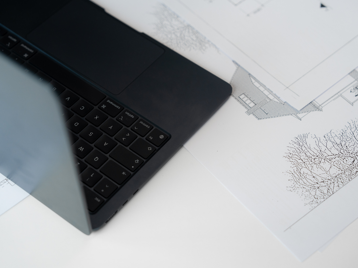 Auf einem weißen Tisch liegen Architekturzeichnungen neben einem geöffneten Laptop.