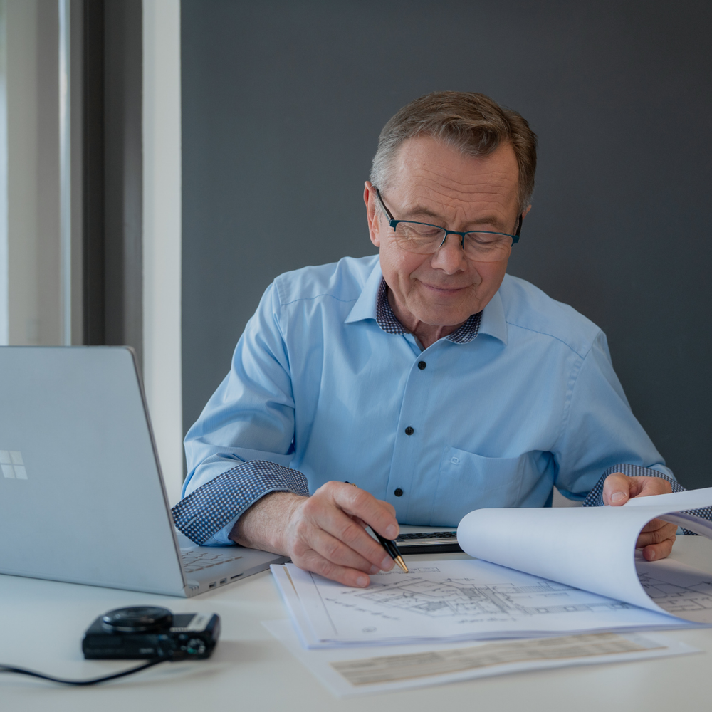 Zertifizierter Sachverständiger Hans-Jürgen Reppe kontrolliert Grundrisspläne am Schreibtisch.